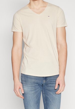 T-shirt beige en col en V en coton doux, avec des manches courtes et un petit logo sur la poitrine. Associé à un jean bleu.