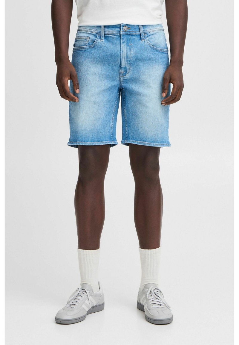 Shorts en denim bleu clair avec une coupe droite, ourlet brut, deux poches avant et fermeture à bouton. Associés à des baskets grises et des chaussettes blanches.