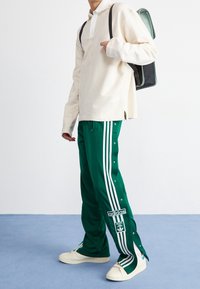 Osoba v zelených sportovních kalhotách Adidas s bílými pruhy a knoflíky, krémovém tričku s dlouhým rukávem, bílých teniskách a nesoucí zelený batoh.