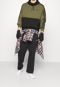 Olivgrön och svart färgblockerad sweatshirt med huva, rutigt skjortband runt midjan, svarta byxor och vita sneakers. Handskar på händerna.