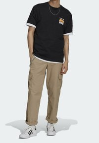 Czarna koszulka z białymi wykończeniami, logo z przodu; beżowe spodnie cargo z bocznymi kieszeniami; białe sneakersy z czarnymi paskami. Srebrny łańcuch jako dodatek.