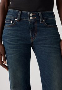 Jean denim bleu foncé avec double boutons argentés et coutures orange, porté avec un haut côtelé noir, montrant la taille et le haut des jambes.
