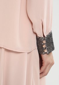 Blusa rosa pallido con maniche lunghe, un polsino nero testurizzato con paillettes e tre bottoni chiari sul polsino.