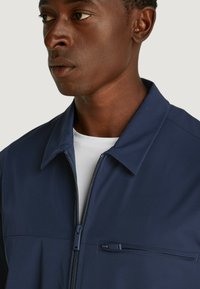 Giacca blu navy con zip e colletto, dotata di tasca sul petto anteriore e texture liscia. Progettata per un aspetto aderente.