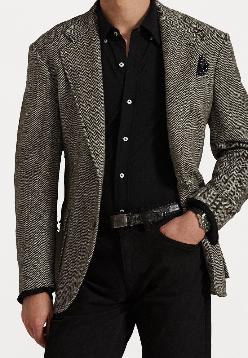 Homme portant un blazer gris en chevrons, une chemise noire boutonnée, un pantalon noir, une ceinture noire et une montre-bracelet, la main dans la poche.