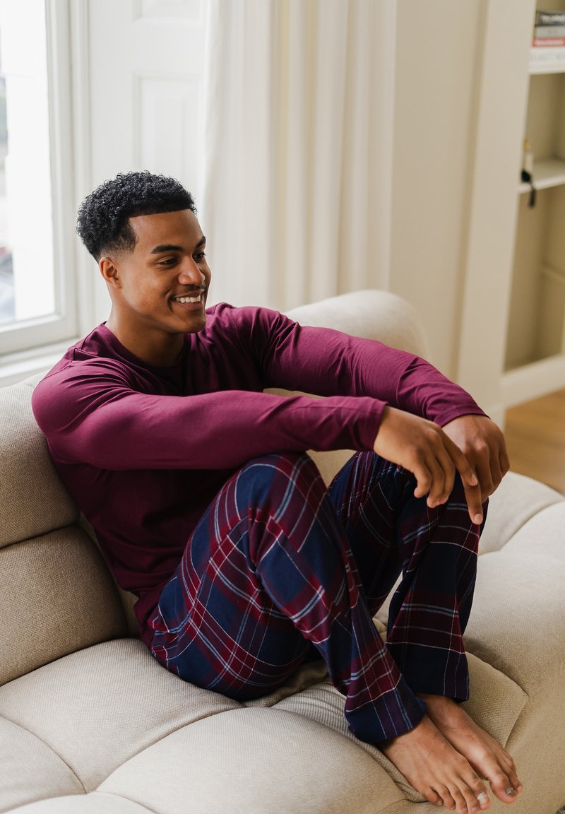 Jonge man in een bordeauxrode langemouwen shirt en geruite pyjamabroek, zittend op blote voeten op een beige bank bij het raam, lachend en ontspannen.
