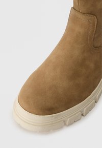 Brązowe zamszowe botki z okrągłym noskiem i teksturowaną powierzchnią. Posiadają białą gumową podeszwę z wyraźnym wzorem bieżnika.