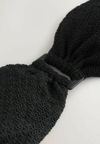 Zwart gestructureerd stoffen accessoire met een geplooid ontwerp en een glanzende zwarte hardwareverbinder in het midden, met een golvend patroon door het geheel.