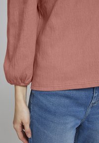Blusa texturizada de color rosa claro con un corte relajado, que presenta mangas abullonadas y un dobladillo que se sitúa justo por encima de la cintura. Lleva puesta con jeans de mezclilla azul.
