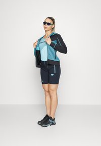 Completo sportivo composto da una giacca leggera azzurra con zip e dettagli neri, una canotta blu coordinata e pantaloni ciclistici blu navy con loghi, abbinati a scarpe nere.