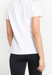 Persona con una camiseta blanca de manga corta y pantalones ajustados negros, vista desde atrás contra un fondo blanco.
