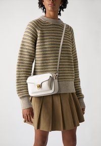 Maglione a righe beige e giallo con polsini a coste, abbinato a una gonna plissettata color khaki. Una piccola borsa a tracolla in pelle bianca presenta dettagli in oro.