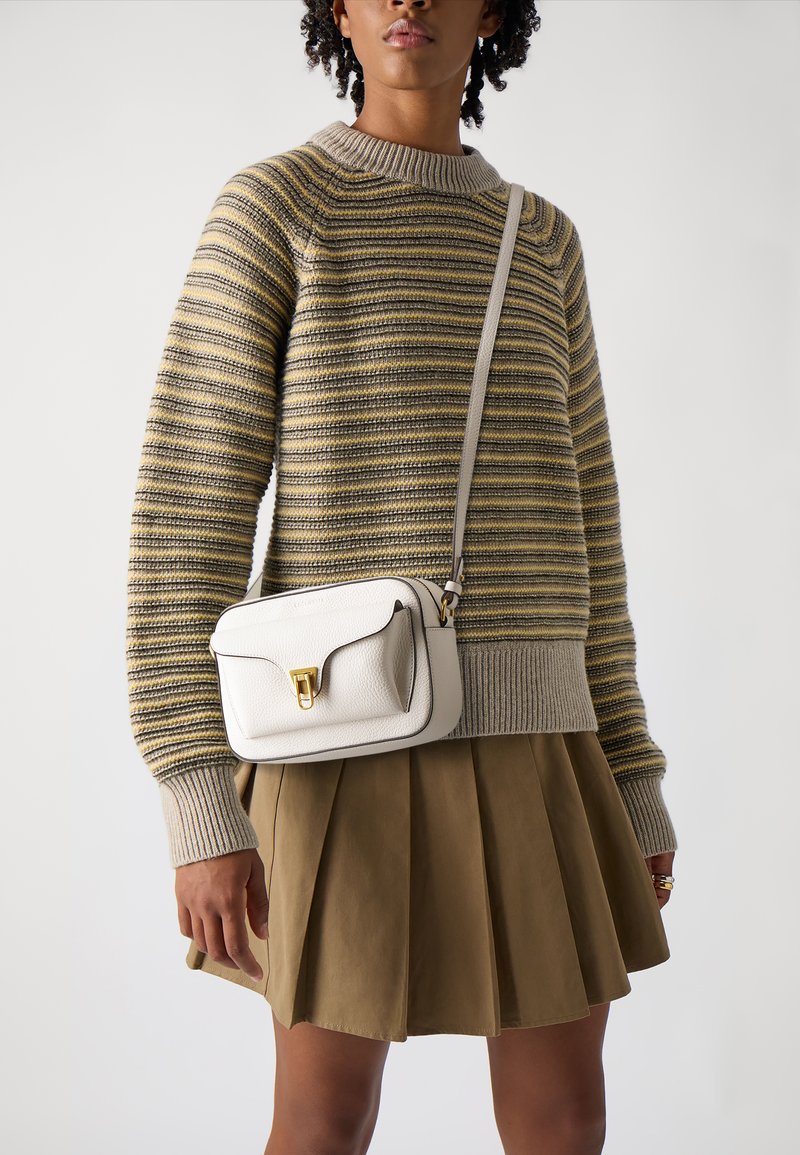 Maglione a righe beige e giallo con polsini a coste, abbinato a una gonna plissettata color khaki. Una piccola borsa a tracolla in pelle bianca presenta dettagli in oro.