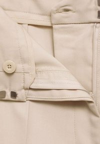 Tissu beige avec un tissage diagonal, présentant un bouton, une poche zippée et des accents métalliques. La texture semble lisse et douce.