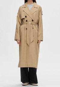 Kvinna klädd i en beige dubbelknäppt trenchcoat med midjeband, svarta vida byxor och beiga skor mot en enfärgad bakgrund.