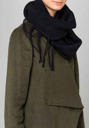 Personne portant un manteau en laine vert olive et une épaisse écharpe noire tricotée avec des franges lâches autour du cou.