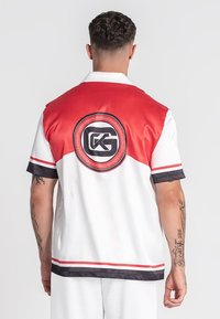 Camisa de manga corta en satén rojo, blanco y negro. Presenta un logotipo circular en la parte posterior y acentos a rayas a lo largo de las mangas y el dobladillo.