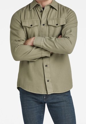 Homme portant une chemise boutonnée vert olive avec des poches poitrine et un jean bleu foncé, bras croisés sur la poitrine sur fond blanc.