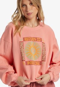 Sweatshirt rose clair à manches longues, arborant un design graphique central d'un soleil et d'un palmier, dans des tons jaunes et terreux.