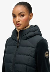 Veste noire matelassée à capuche avec fermeture à glissière et accents en tissu texturé. Dispose d'un écusson de marque sur le bras gauche. Forme lisse et contemporaine.