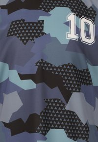 Sininen kamouflagepaita, jossa on geometrinen kolmionmuotoinen kuvio mustana ja vaaleampina sävyinä, etuosassa valkoinen numero "10".