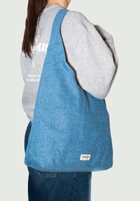 Sac fourre-tout en denim bleu avec des bretelles larges et une forme carrée, doté d'une petite étiquette blanche sur la couture du bas, en tissu texturé, sans quincaillerie visible.