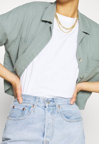 Camicetta verde chiaro a maniche corte sopra una maglietta bianca, abbinata a jeans a vita alta di colore blu chiaro, con bottoni e tasche.