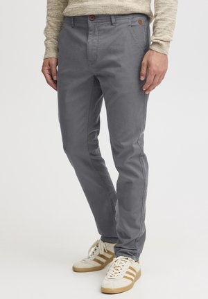 Mężczyzna ubrany w dopasowane szare spodnie, beżowy sweter dziany oraz białe tenisówki z brązowymi paskami, stojący na jednolitym tle.