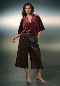 Top en velours bordeaux avec un col en V et des manches larges associé à un pantalon large en similicuir marron. Le mannequin porte des talons dorés à lanières.
