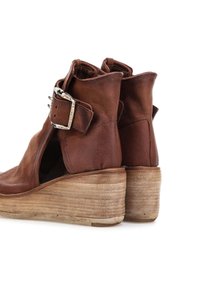 Brune læder ankelstøvler med kilehæle, sølv spænder og strukturerede stablede træhæle, set fra en vinkel på hvid baggrund.