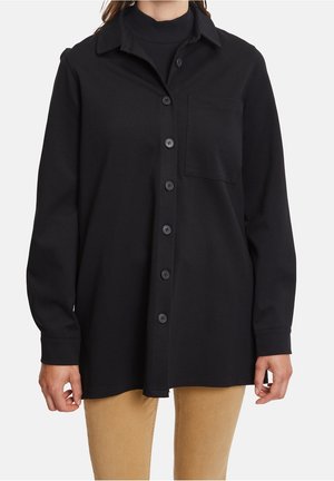 Mujer con camisa negra de botones y bolsillo en el pecho, encima de un suéter de cuello alto negro, combinada con pantalones marrón claro.