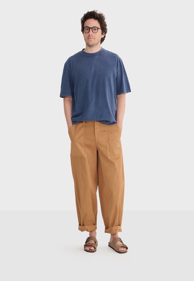 Homme aux cheveux bouclés et portant des lunettes, vêtu d'un t-shirt bleu ample, d'un pantalon couleur sable retroussé, et de sandales marron, debout les mains dans les poches.