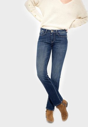 Osoba ubrana w dopasowane ciemnoniebieskie dżinsy, brązowe botki do kostki oraz kremowy sweter dziany z złotymi guzikami na rękawach.