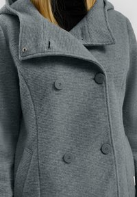 Manteau double boutonnage gris texturé avec quatre boutons, porté sur un col roulé noir, montré des épaules jusqu'au milieu du torse.