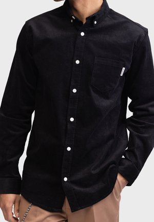 Man draagt een zwarte corduroy overhemd met witte knopen en borstzak, gecombineerd met lichtbruine broek en kettingaccessoire.