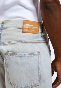 Jasnoniebieskie dżinsy denimowe z prostokątną brązową skórzaną łatką z napisem "DRDENIM" oraz informacjami o pielęgnacji. Widoczne szwy kieszeni tylnych.