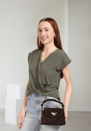 Sac à main en daim marron avec un emblème d'abeille doré et une bandoulière chaîne. Le modèle porte un haut vert olive au design drapé et un jean bleu clair.