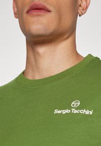 Zöld pamut póló kerek nyakkivágással, fehér "Sergio Tacchini" logóval és egy kör alakú emblémával a bal mellrészen.