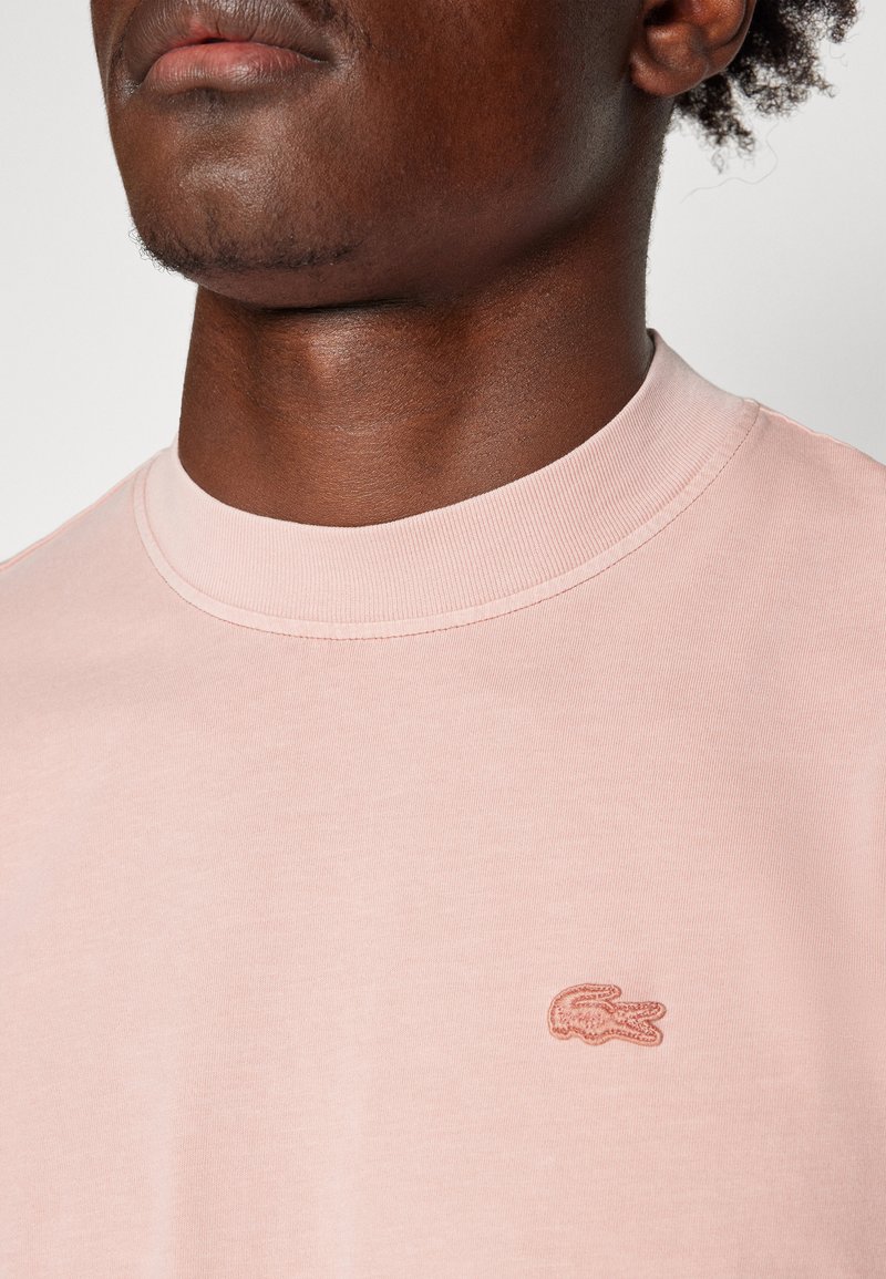 T-shirt en coton rose clair avec un col rond et un col côtelé. Présente un logo brodé sur la zone de la poitrine gauche. Texture lisse.