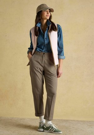 Giovane donna con cappellino beige, gilet rosa chiaro sopra camicia azzurra abbottonata, pantaloni a quadri e sneakers verdi, in piedi con le mani in tasca.