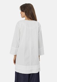 Witte katoenen tuniek met een losse pasvorm, driekwart mouwen en oogjesdetails bij de mouwen. Heeft een ronde halslijn en een rechte achterkant.