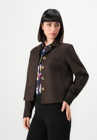 Žena u tamnosmeđem sako s dugmadi preko šarene cvjetne majice i crnih hlača, pozira s jednom rukom na bokovima na bijeloj pozadini.