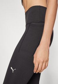 Leggings noires taille haute en tissu lisse et extensible avec des coutures plates. Présente un petit logo blanc Puma sur la cuisse gauche.