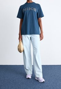 Osoba oblečená v modrém tričku Nike, světlých modrých džínech a růžových teniskách Nike, držící malou béžovou psaníčko, stojící na modrém koberci.