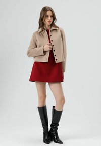 Beige cropped jas met kraag, zwarte knopen, gecombineerd met een rode jurk en zwarte leren laarzen tot aan de knie. Soepele textuur van de stof.