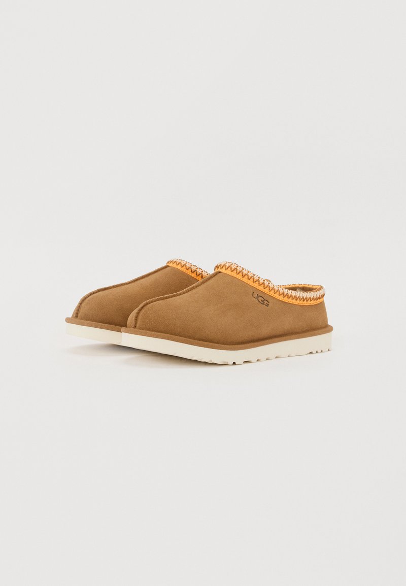 Pantofole in suede marrone con rifiniture intrecciate arancioni nella parte superiore, dotate di una suola in gomma bianca, punta tonda e design minimalista.