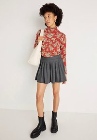 Rotes langärmeliges Oberteil mit Blumenmuster, grauer plissierter Rock mit Nadelstreifen, schwarze Plateau-Stiefeletten und eine cremefarbene Schultertasche.