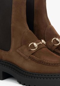 Brązowe zamszowe botki za kostkę z czarnymi elastycznymi bocznymi panelami, złotymi akcentami ozdobnymi oraz grubą czarną gumową podeszwą z teksturowanym bieżnikiem.