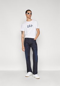 Hvid t-shirt med "GAP" logo, mørkeblå jeans og hvide sneakers. Modellen har hvide solbriller på og står foran en neutral baggrund.
