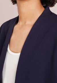 Gros plan d'une personne portant un blazer bleu marine sur un haut blanc, montrant le cou et la partie supérieure de la poitrine avec un teint naturel.